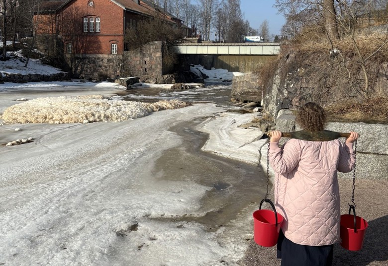 Nainen kantaa kahta ämpäriä puisella kantotelineellä harteillaan. Taustalla näkyy Vantaanjoki ja punainen tiilirakennus. 