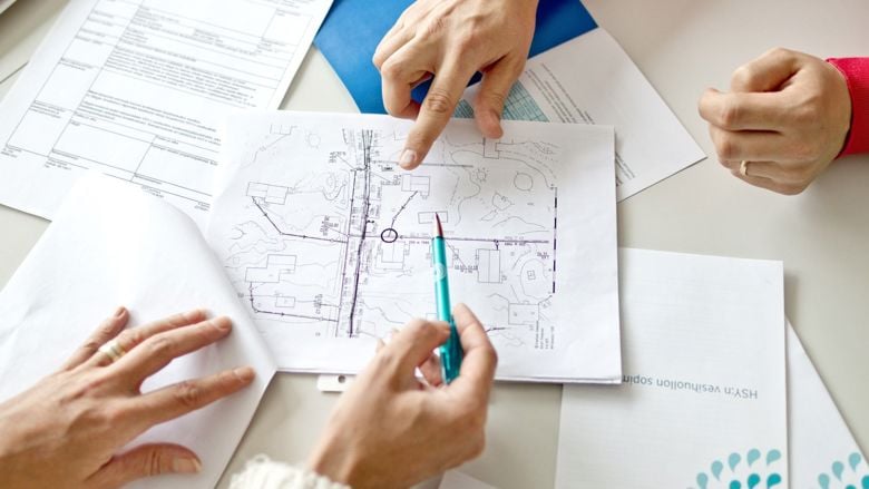 Pöydällä on papereita. Papereita tutkivien henkilöiden kädet näkyvät ja yksi osoittaa paperia.