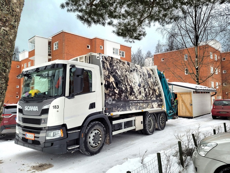 Taidejäteauto kuvattuna talvella kahden kerrostalon edessä jätetilan. Auton kyljessä teksti: Lasit ja metallit ovat ikuisia - kiitos kun lajittelet.