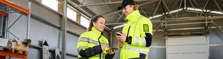 Two workers in high-visibility yellow jackets with reflective strips inside a warehouse, smiling and looking at a mobile device.